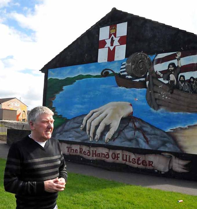 Taxifahrerkollege Bobby Thompson vor einer Hauswand mit einer roten Hand, der „Red Hand Ulster“, die auch auf der Flagge von Nordirland zu sehen ist.
