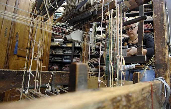 Eine junge Frau sitzt fast versteckt zwischen den labyrinthartigen Regalen und webt eine schmale goldene Borte. Eine junge Frau sitzt fast versteckt zwischen den labyrinthartigen Regalen und webt eine schmale goldene Borte.