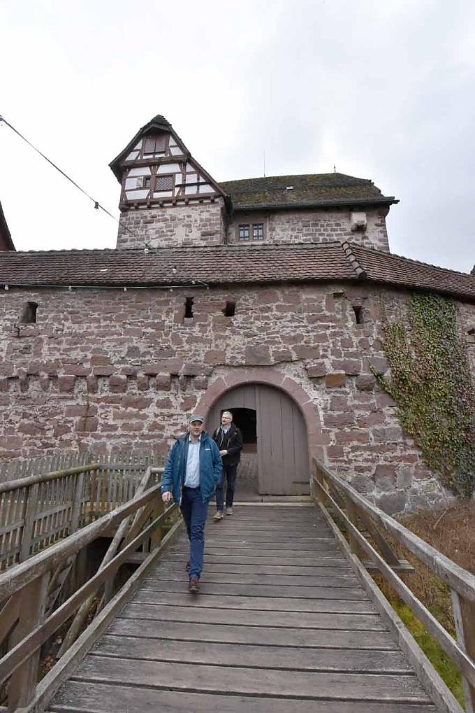Besucher verlassen das Schloss über die Holzbrücke. Familie in einer Fensternische mit Sitzbänken.